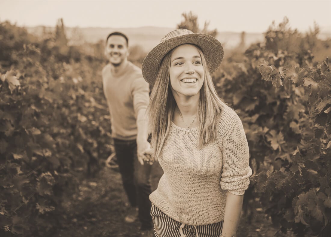 A sepia photo of a lady leading a man by the hand through a vineyard.