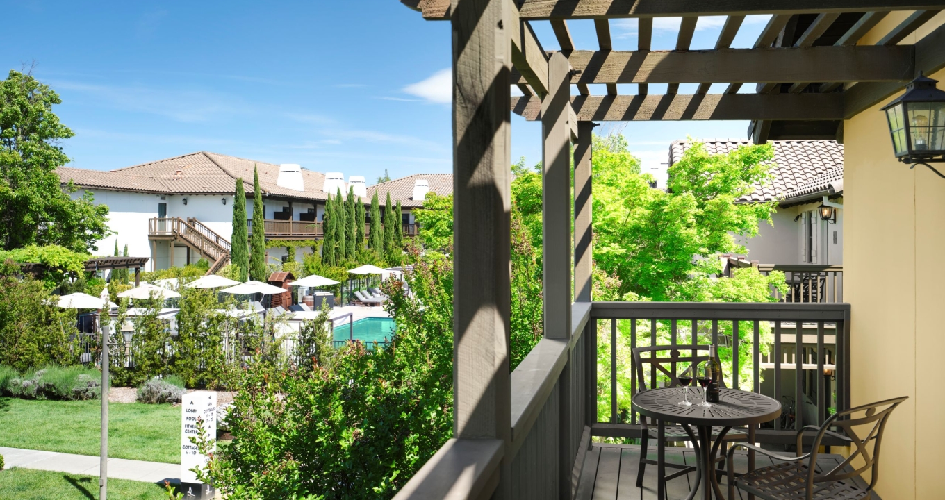 Balcony seating area with a round metal table and chairs under a wooden pergola, overlooking a sunny courtyard with trees, umbrellas, and townhouses.