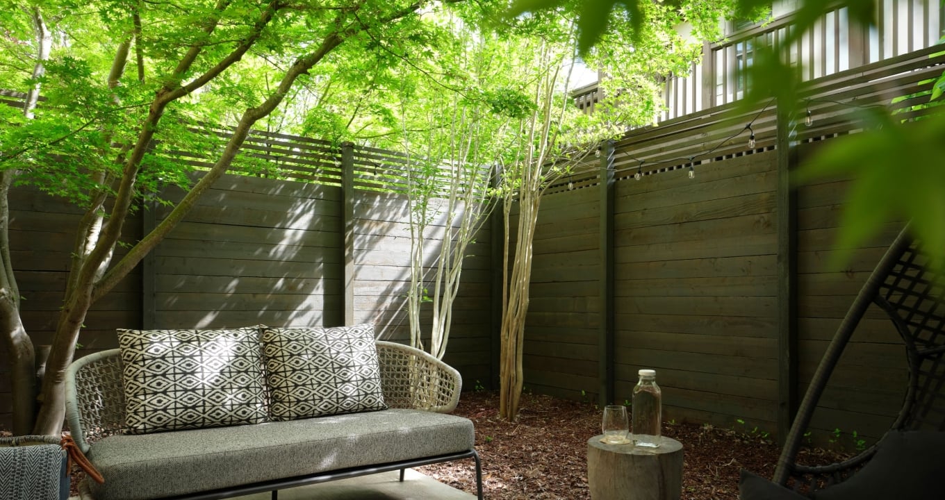 Outdoor seating area with a gray cushioned wicker sofa and geometric-pattern pillows under lush green trees beside a wooden privacy fence.