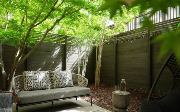 Outdoor seating area with a gray cushioned wicker sofa and geometric-pattern pillows under lush green trees beside a wooden privacy fence.
