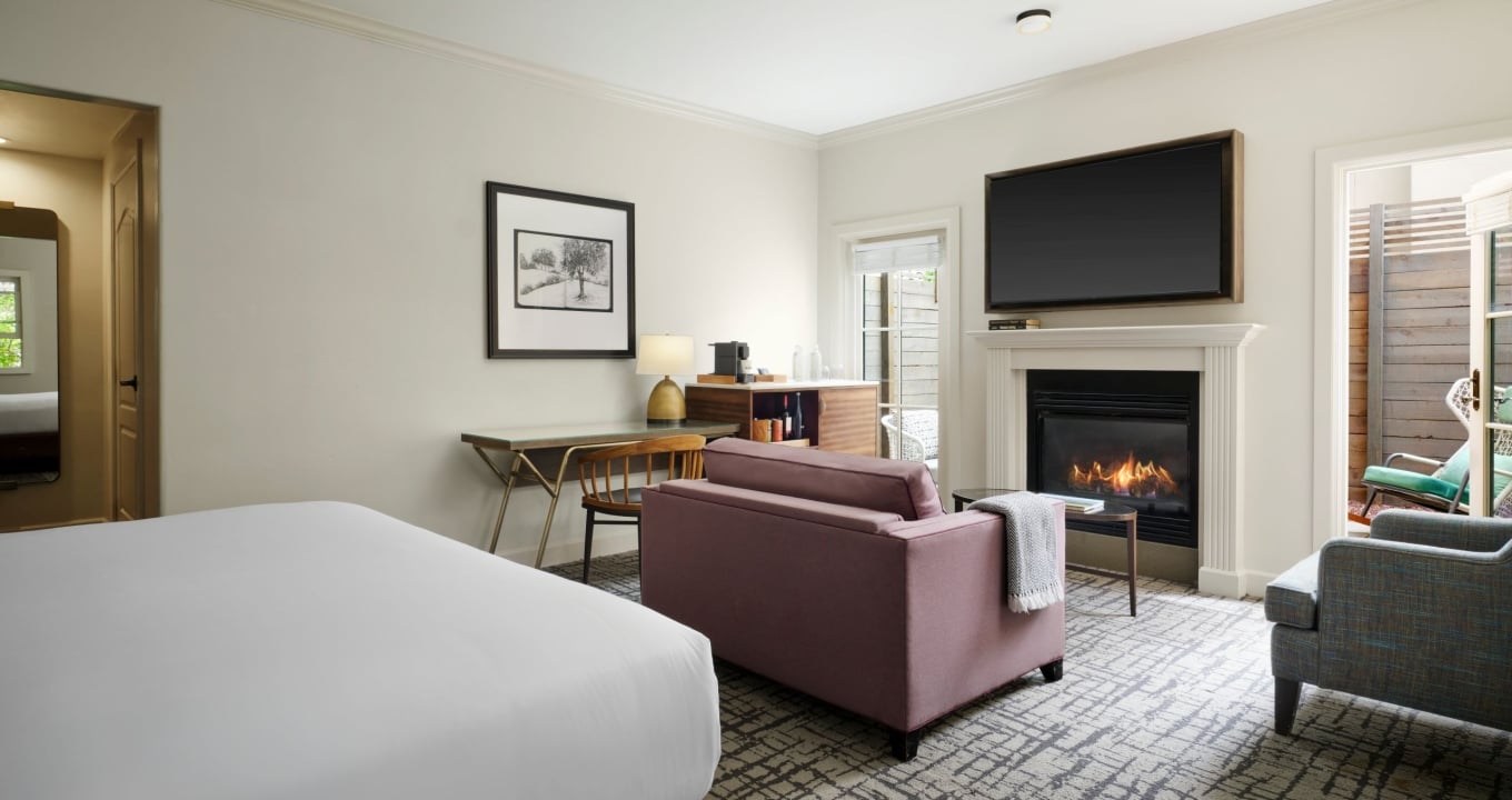 Hotel suite living area with a bed in the foreground, pink sofa, fireplace, and wall-mounted TV above the mantel.