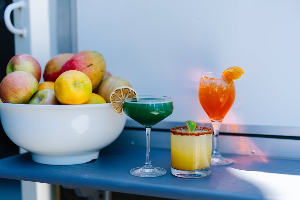 Three craft cocktails displayed with fresh apples and citrus in a white bowl on a blue table at sunset, creating an elegant luxury hospitality scene.