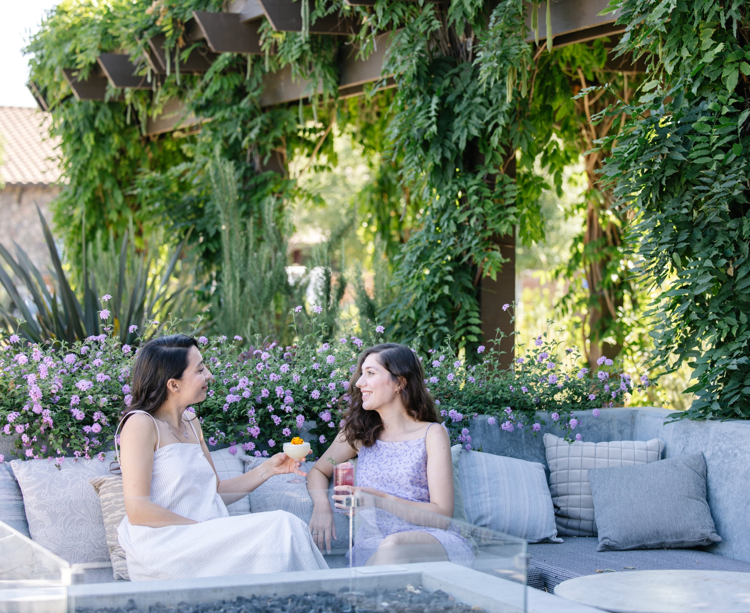Two women in light dresses sit on a gray outdoor sofa, chatting and holding drinks, with pink flowers and green ivy surrounding them.