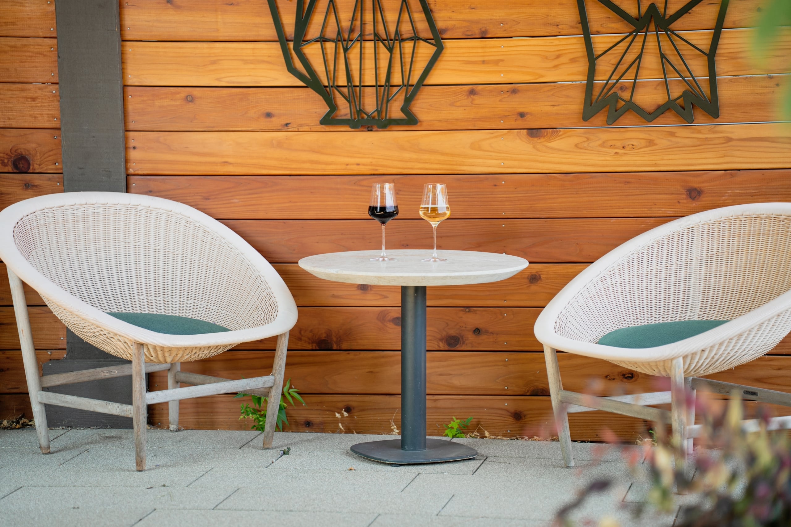 Two wicker chairs flank a small round table with two wine glasses outdoors against a wooden paneled wall.