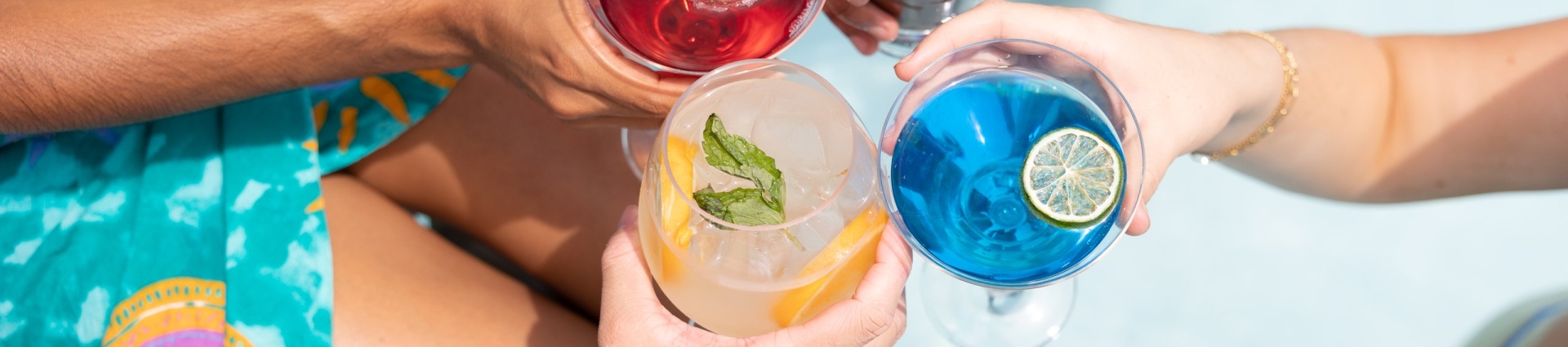 Four people holding colorful cocktails by a pool, toasting together in bright daylight wearing vibrant swimwear.