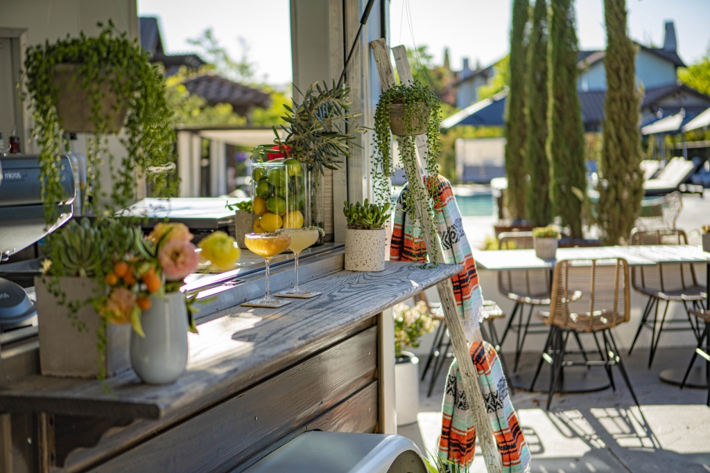 Poolside bar with cocktails, hanging succulents, and vibrant textiles overlooking a sunny resort pool and cypress trees.