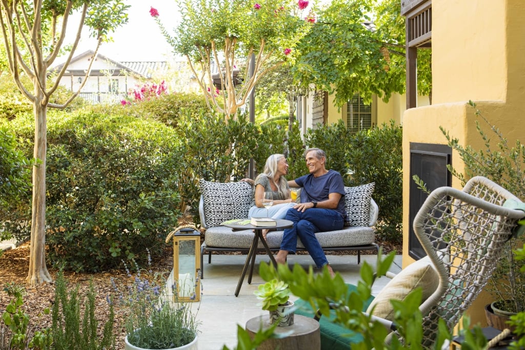 Couple relaxing on outdoor sofa in lush garden courtyard with flowering trees and contemporary furniture.