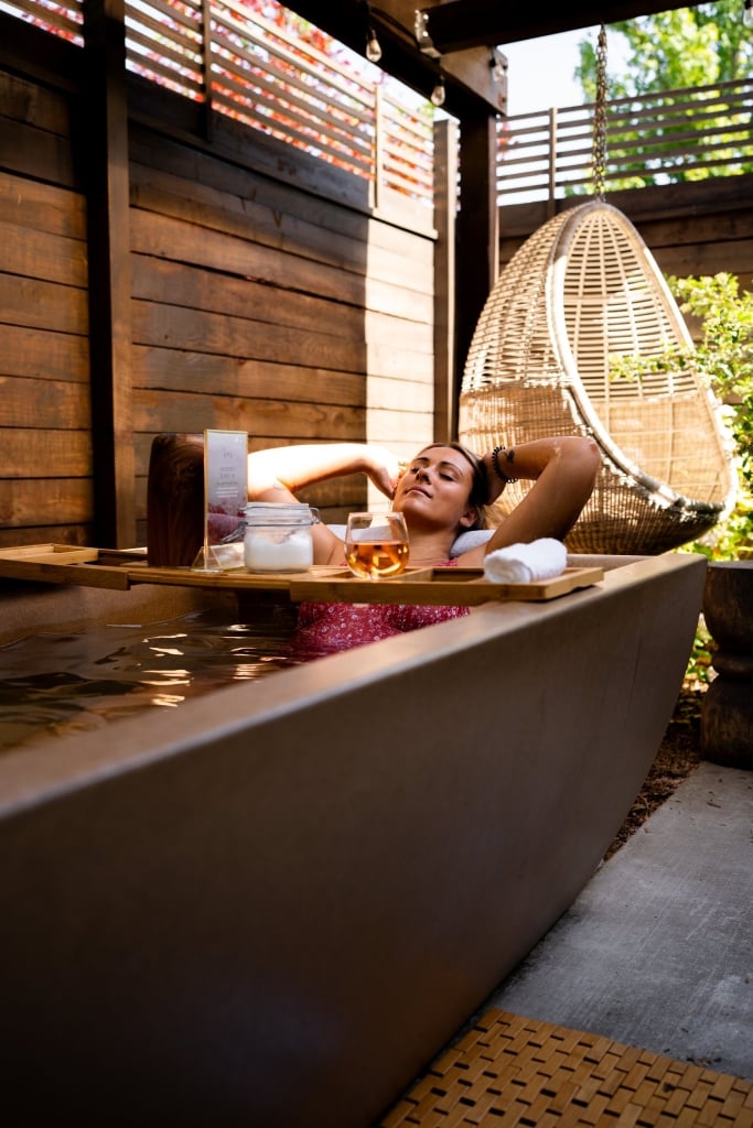 Woman relaxing in outdoor spa tub with suspended chair, spa products, and natural wood architecture in warm golden light.