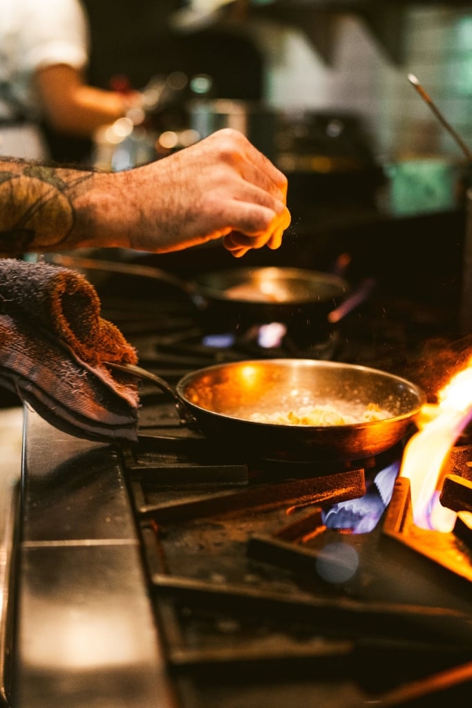 Chef in dark apron cooking over open flame in professional kitchen with warm golden lighting