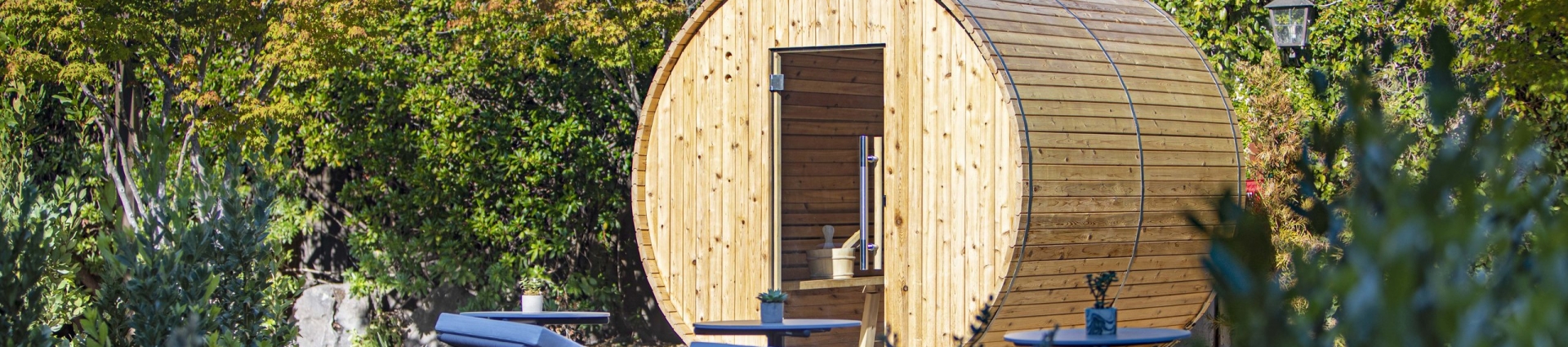 Circular wooden barrel sauna in a lush garden with lounge chairs and flowering shrubs nearby.