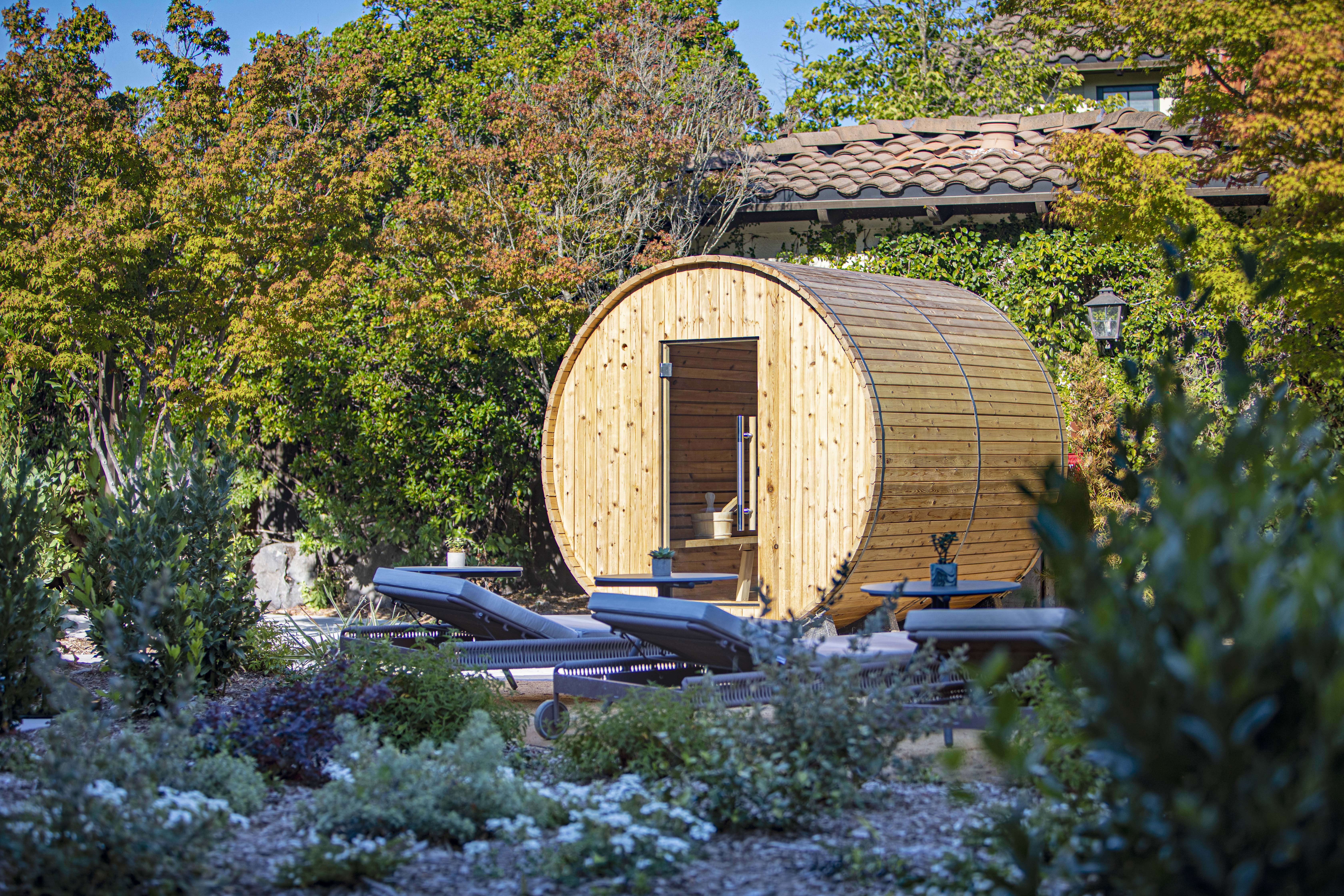 Circular wooden barrel sauna in a leafy backyard with lounge chairs nearby.