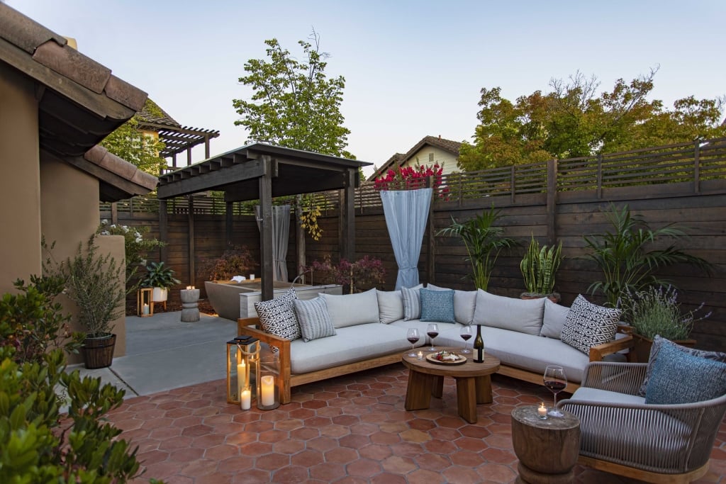 Luxurious outdoor patio with L-shaped sectional sofa, wooden coffee table, and candles surrounded by dark wood fencing and flowering plants at dusk.