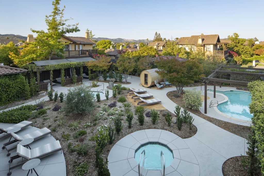 Aerial view of luxury spa grounds with hot tub, pool, barrel sauna, pergola cabanas, and landscaped gardens.