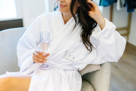 Woman in a white spa robe seated on a gray chair, holding a pink champagne flute.