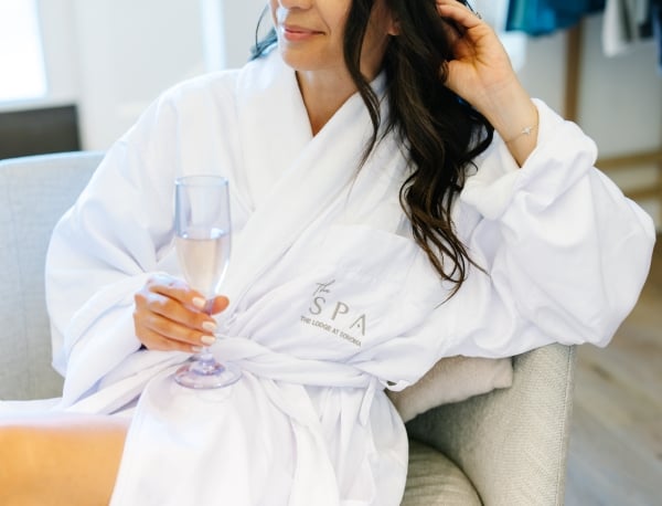 Woman in a white spa robe seated on a gray chair, holding a pink champagne flute.