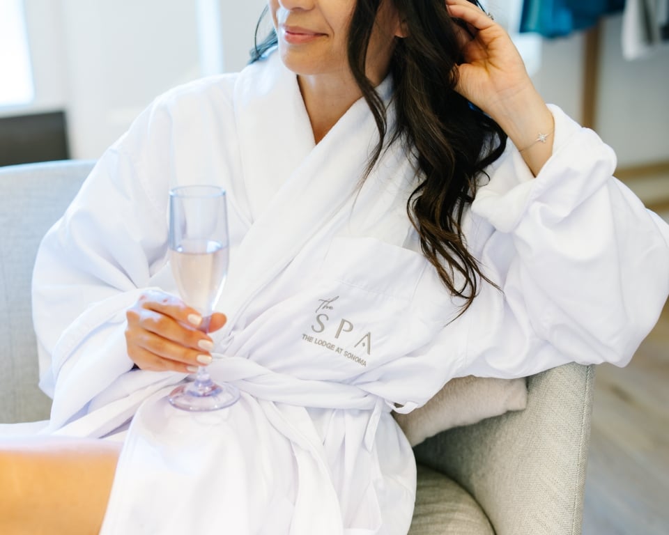 Woman in a white spa robe seated on a gray chair, holding a pink champagne flute.