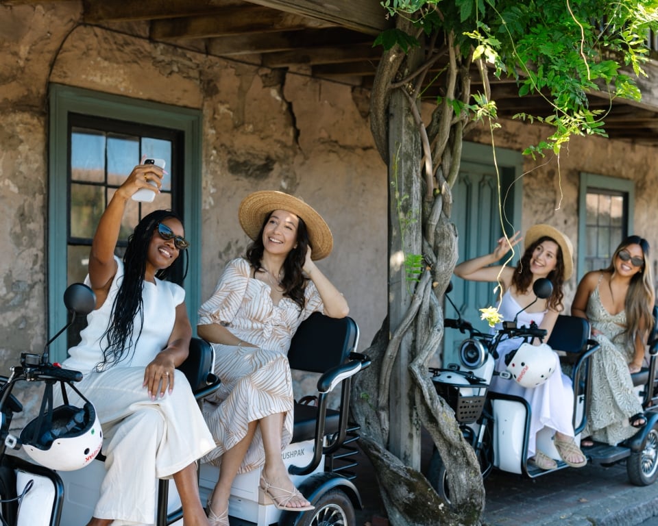 Four women on white electric scooters posing for a group selfie in front of a rustic wall with ivy and greenery above.