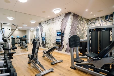 Indoor gym with cardio machines (treadmills and ellipticals) on a wooden floor and a tree mural along the wall.