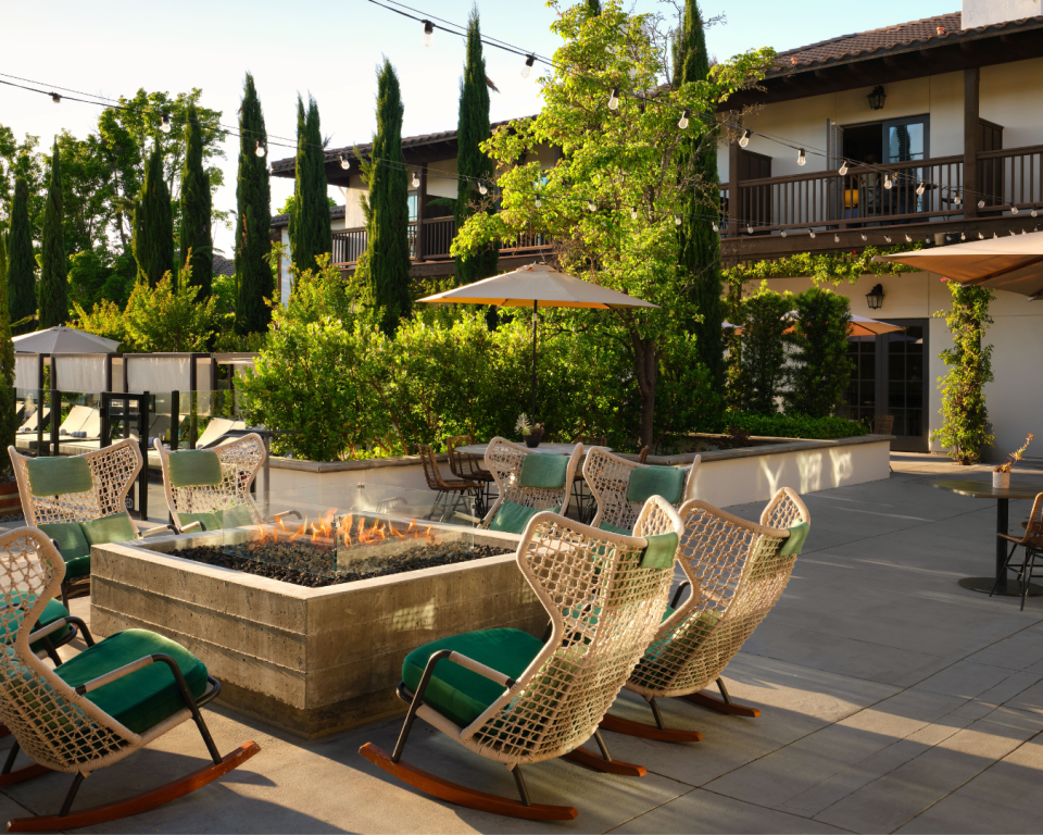 Outdoor hotel courtyard with a rectangular fire pit, wicker chairs with green cushions, string lights, and umbrellas under a two-story building.