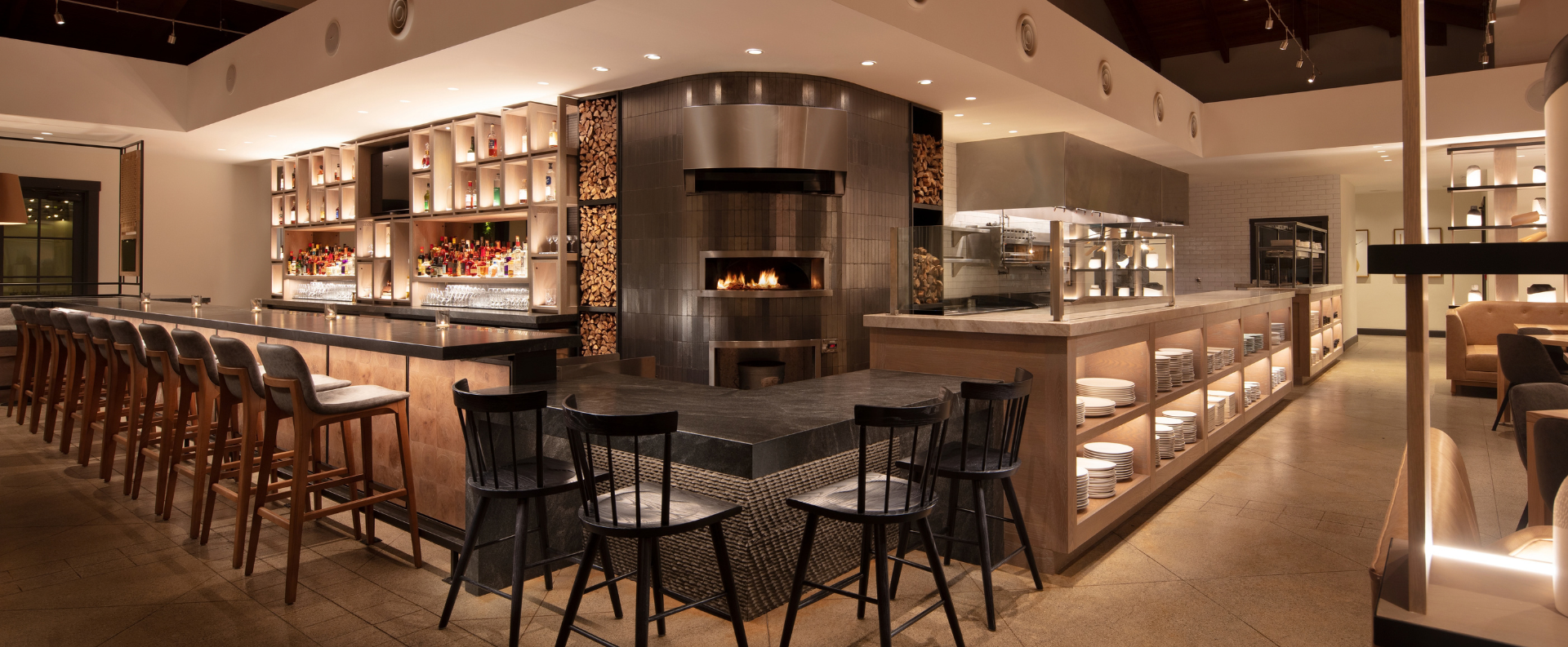Luxurious bar with a long counter, wooden stools, backlit liquor shelves, and a central tiled oven area.