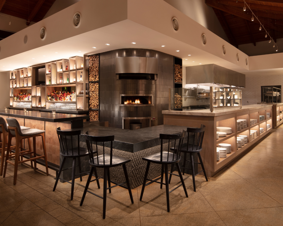 Luxurious bar with a long counter, wooden stools, backlit liquor shelves, and a central tiled oven area.