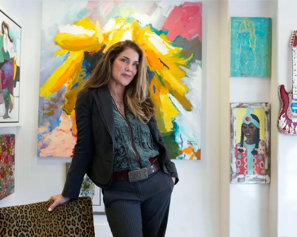 Woman in a dark pinstripe suit leaning on a leopard-print chair in an art gallery with bright abstract paintings behind her.
